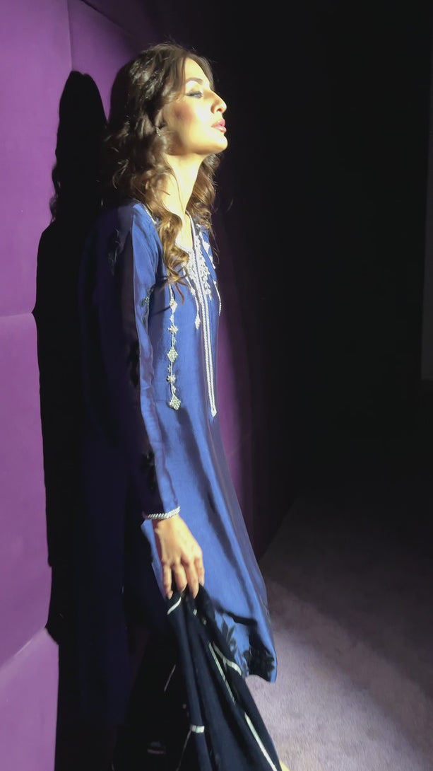 A woman models a regal royal blue velvet kameez featuring silver tilla embroidery and geometric motifs, paired with slim-fit trousers and a matching embroidered velvet shawl.


